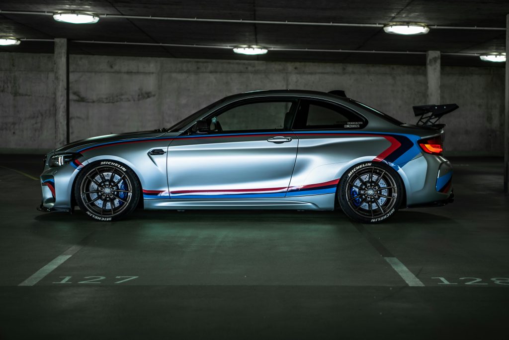 pexels-photo-12249451-12249451 Side view of a BMW M2 sports car with racing stripes, parked in a dimly lit garage.
